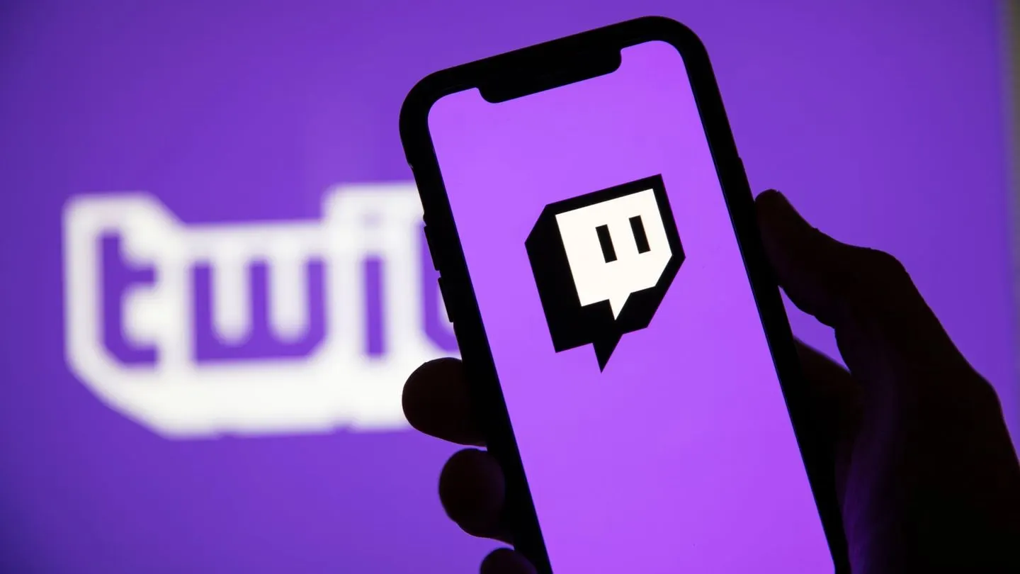 A hand holding a phone showing the Twitch logo, with the Twitch wordmark on a purple screen behind it